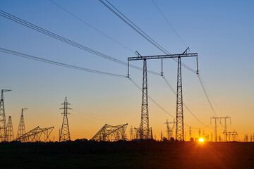 High voltage electricity towers in field at sunset and clear blue sky. Dark silhouettes of repeating power lines on orange sunrise. Electricity generation, transmission, and distribution network