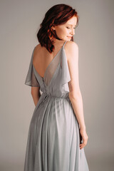 Fototapeta premium Beautiful young woman in dust blue dress. Studio portrait of ginger lady in evening gown dancing on high heel shoes in studio. Bridesmaid's outfit for hen party, rear view.