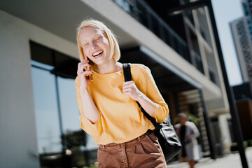 Fototapeta premium Happy girl walking on the street using the phone. Beautiful blonde girl talking to the smartphone.