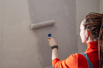 Woman with dreadlocks, in an orange turtleneck and denim jumpsuit is painting the wall with a roller in gray color. Concept of repair, renovation of the new apartment