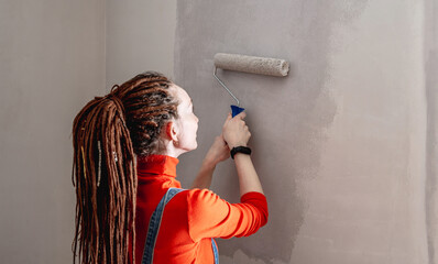 Woman with dreadlocks, in an orange turtleneck and denim jumpsuit is painting the wall with a roller in gray color. Concept of repair, renovation of the new apartment