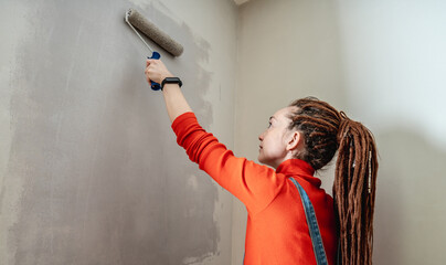 Woman with dreadlocks, in an orange turtleneck and denim jumpsuit is painting the wall with a roller in gray color. Concept of repair, renovation of the new apartment