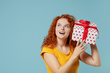 Young happy redhead woman wear yellow t-shirt hold red present box with gift ribbon bow look aside on workspace area isolated on plain light pastel blue background studio. People lifestyle concept