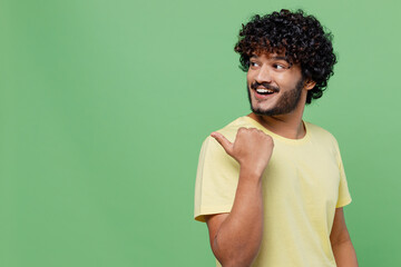 Young smiling happy Indian man 20s in basic yellow t-shirt point thumb finger aside on workspace area mock up isolated on plain pastel light green background studio portrait. People lifestyle concept.