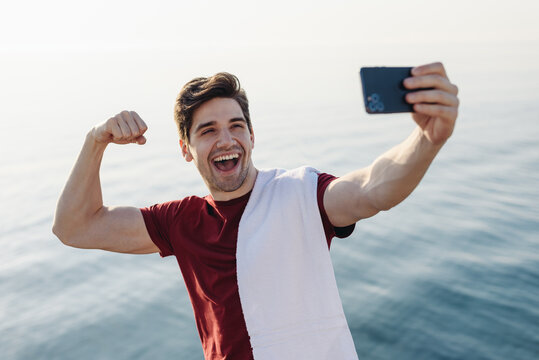 Young strong sporty athletic fit sportsman man in sports clothes do selfie shot mobile cell phone show muscles warm up training at sunrise sun dawn over sea beach outdoor on pier seaside in morning.