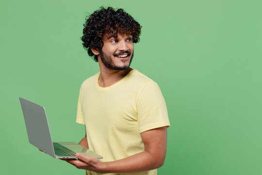 Young happy Indian man 20s in basic yellow t-shirt hold use work on laptop pc computer look aside on workspace isolated on plain pastel light green background studio portrait People lifestyle concept