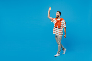 Fototapeta na wymiar Full body side profile view young smiling caucasian cheerful man 20s wear orange striped t-shirt walking going waving hand isolated on plain blue background studio portrait. People lifestyle concept.