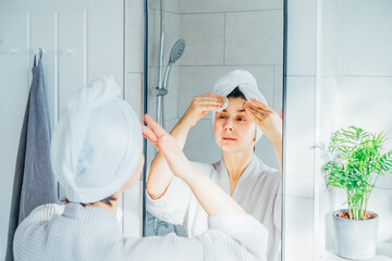 Woman in bathrobe with a towel on head looking in mirror, exfoliating facial skin with loofah...