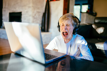 Blond child boy in headphones is using a laptop and study with video call teacher at home. Pupil...