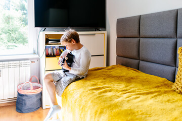 Cute blonde caucasian boy sitting on sofa playing acoustic guitar while practicing in his room.