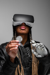 Fototapeta premium KYIV, UKRAINE - APRIL 28, 2022: Blurred african american woman in vr headset with robotic hand holding bitcoin isolated on grey.