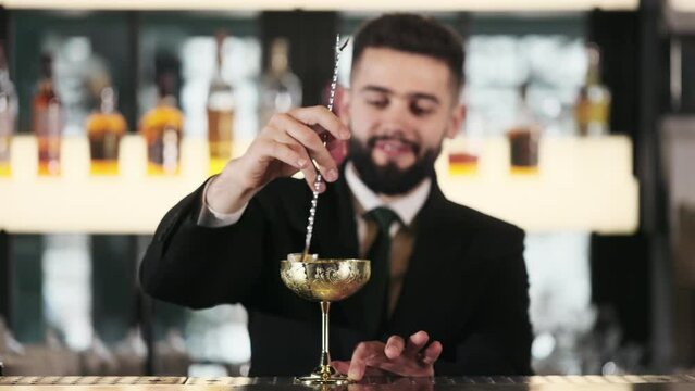 Caucasian man making exclusive alcoholic cocktail at bar counter using golden vintage glass, spoon and various ingredients. Skillful handsome bartender in suit stirring drink in unique shiny goblet.
