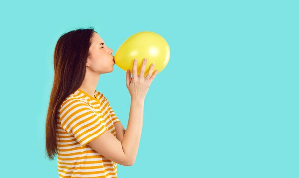 Young woman preparing for party funny blowing yellow balloon isolated on light blue background. Profile portrait of girl in casual T-shirt who with funny expression inflates balloon near copy space.