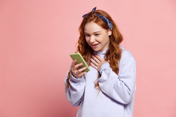 Surprised young redhead girl, student using phone isolated on pink studio background. Human emotions, facial expression concept. Trendy colors