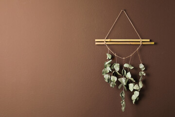 Beautiful decor element made of dry bamboo sticks and eucalyptus branches hanging on brown wall, space for text