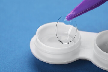 Taking contact lens from case with tweezers on blue background, closeup