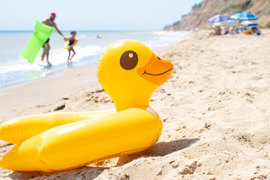 Yellow inflatable duck ring laying on sandy beach near blue wavy ocean in sunny day with people and sun umbrellas on background. Protection swim tube for kid. Summer travel vacation resort concept