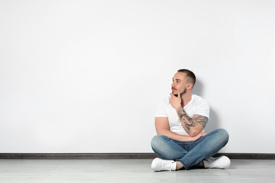 Handsome young man sitting on floor near white wall indoors, space for text