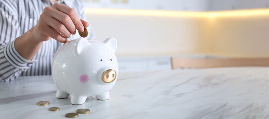 Woman putting coin into piggy bank at white marble table indoors, closeup view with space for text. Banner design