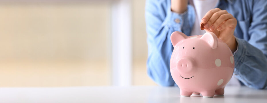 Woman putting coin into piggy bank at table indoors, closeup view with space for text. Banner design