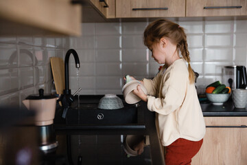 A small child with a rag in his hands stands on a chair near the sink and washes dishes. A little girl helps her mother with cooking and cleaning in the kitchen