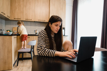 Freelancer woman sits by the table in the home kitchen office, working on laptop. Playful child distracts from work, kid making noise and asking attention from busy mom