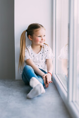 Beautiful little girl smiling and watching out the window. A child looks out the window. Young girl looking from window at windowsill.