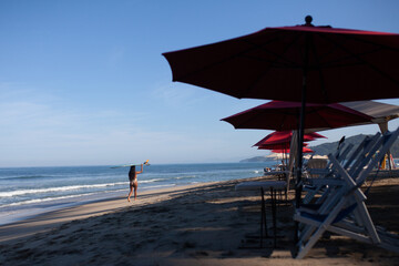 Mujer caminando con tabla de surf en la arena, sombrillas rojas y el cielo azul, playa de Sayulita, Jalisco, México, mujer surfista en traje de baño, mar y sol, deporte acuático