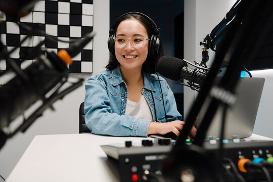 Beautiful happy young female radio host using microphone and laptop while broadcasting in studio