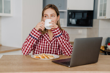 Young white happy woman having breakfast and using laptop in kitchen