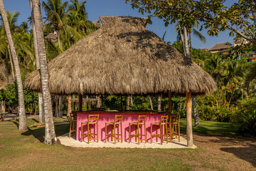 Fototapeta na wymiar Palapa de playa con barra bar de color rosa en México bajo el sol, con palmeras y cielo azul, bancas rusticas tejidas de palma y jardín