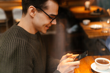 Young man smiling while drinking coffee and using cellphone in cafe