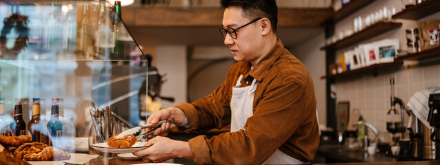 Asian man holding plate with croissant standing at counter in cafe