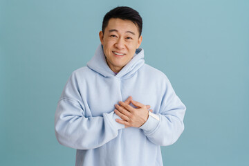 Brunette adult asian man smiling and keeping hands on his heart