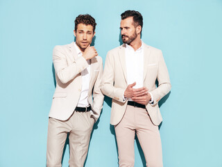 Portrait of two handsome confident stylish hipster lambersexual models. Sexy modern men dressed in white elegant suit. Fashion male posing in studio near blue wall