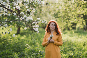 Fototapeta na wymiar Portrait of gorgeous middle aged woman in casual dress relaxing in a blooming spring garden. Happy senior woman smiling and looking at camera. Red-haired mature lady posing outdoors in a spring day.