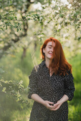 Portrait of gorgeous middle aged woman in casual dress relaxing in a blooming spring garden. Happy senior woman smiling and looking at camera. Red-haired mature lady posing outdoors in a spring day.