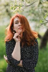 Fototapeta premium Portrait of gorgeous middle aged woman in casual dress relaxing in a blooming spring garden. Happy senior woman smiling and looking at camera. Red-haired mature lady posing outdoors in a spring day.