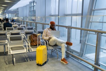 Pensive African man sitting in hall of airport terminal using laptop, wear headphones and listening music. Thoughtful passenger travelling with backpack, online working on computer and waiting landing