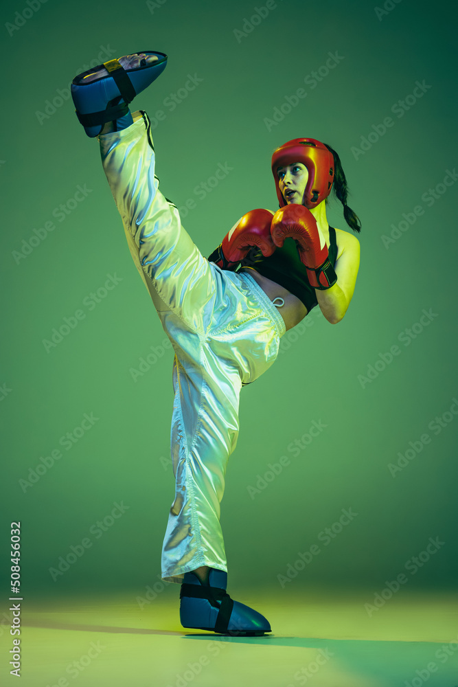 Wall mural Professional female boxer in boxing gloves and helmet in action, motion isolated on green studio background in neon light. Sport, competition, challenges, results, success concept