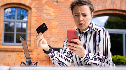 Upset young caucasian woman using online banking, problem with blocked credit card, looking confused at smartphone sitting on red brick building background. Checking balance, internet fraud concept.