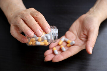 Wrinkled hands of elderly woman with pills on dark wooden table background. Medication in capsules,...