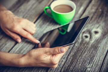 Tablet computer in hands on the background of a wooden table and a cup with a drink.