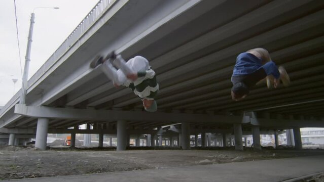 Two professional parkourists running towards each other, then performing cartwheels and backflips while training together outdoors in city
