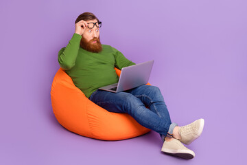 Full length of impressed handsome man sit bag hand touch glasses isolated on violet color background