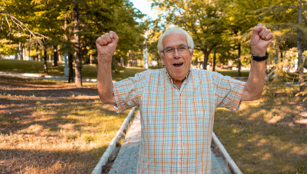 Hombre mayor en el parque, con gesto de victoria, con los brazos levantados y expresión alegre. Fotografía horizontal con espacio para texto.