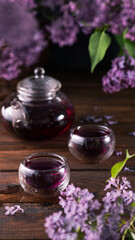 Obraz na płótnie Canvas Purple tea is poured from a glass teapot into cups. Lilac flowers are all around