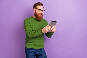 Photo of intelligent focused young male in spectacles searching information in tablet isolated on purple color background