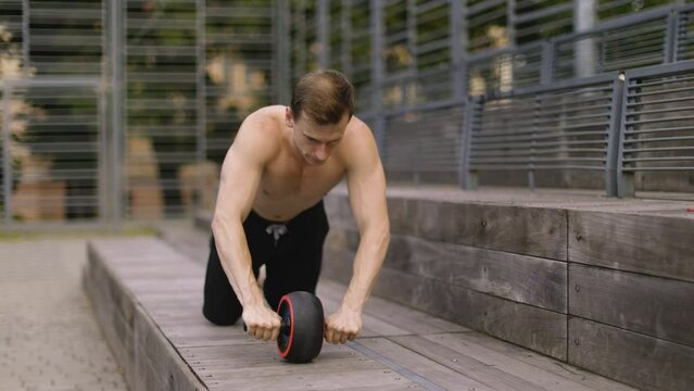 Athletic sporty caucasian shirtless man doing exercise with abs roller wheel to strengthen his abdominal muscle in outdoor gym.