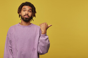 Studio portrait of young african american man posing over studio background points with a finger aside at copy space with emotionless facial expression